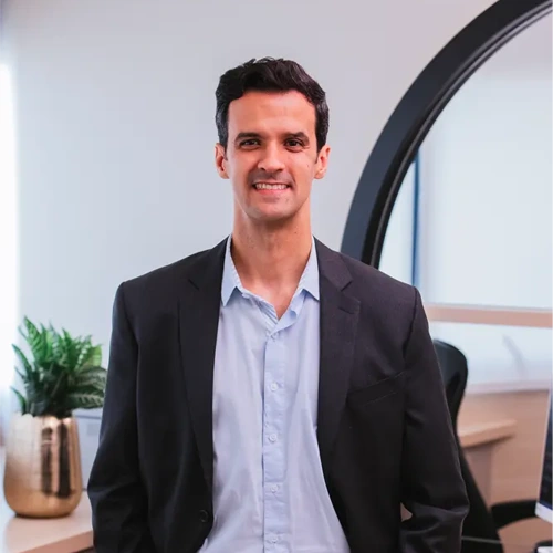 Retrato profissional do Dr. Marcelo de Loyola e Silva Avellar Fonseca, reumatologista da Clínica Loyola & Avellar, em Curitiba. Ele está em pé, sorrindo, vestindo um paletó preto sobre uma camisa social azul clara. O ambiente é um consultório moderno e bem iluminado.