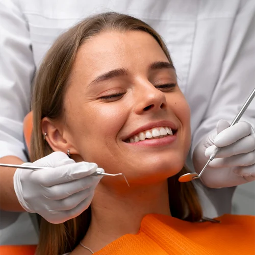 Close-up de uma paciente jovem sorrindo com os olhos fechados durante um procedimento odontológico preventivo na Loyola & Avellar. Um dentista usando luvas brancas utiliza instrumentos de precisão, como um espelho clínico e uma sonda, para realizar o check-up. A paciente está acomodada em uma cadeira com protetor laranja vibrante.