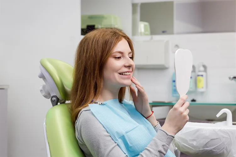 Close-up lateral de uma paciente ruiva sorrindo e tocando o queixo enquanto se olha em um espelho de mão branco, sentada em uma cadeira odontológica verde menta em um consultório dentário moderno e bem iluminado.