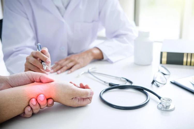 Close-up em ângulo baixo focado nas mãos de um paciente sobre uma mesa branca de consultório médico. O médico, vestindo jaleco branco e segurando uma caneta, observa enquanto a região do punho e articulações da mão do paciente apresenta um destaque gráfico vermelho brilhante, simbolizando dor aguda e inflamação. Um estetoscópio e frascos de medicamentos estão visíveis ao fundo, fora de foco.
